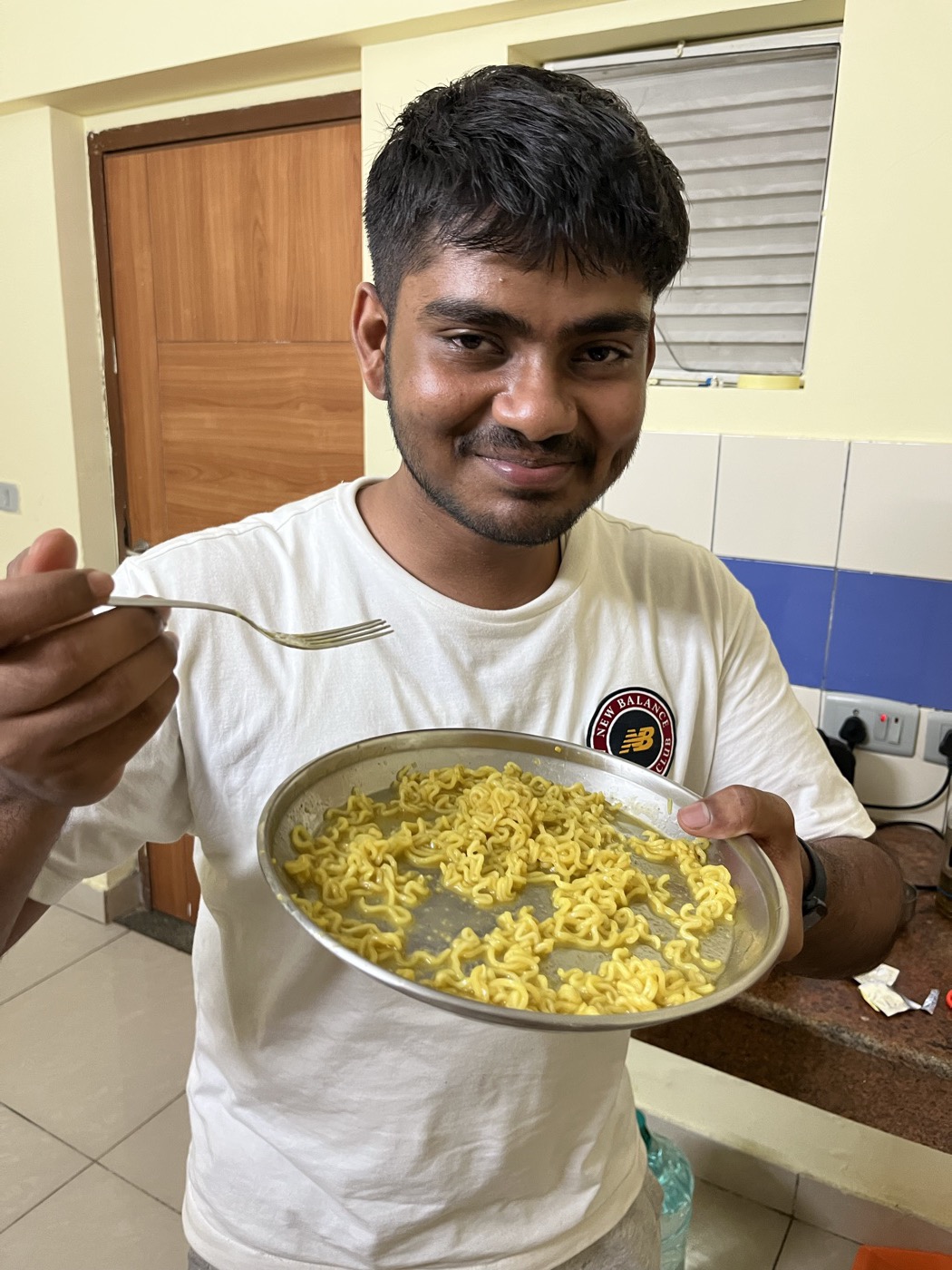 A friend smiling at the camera, holding a plate of noodles in one hand and a fork in the other.